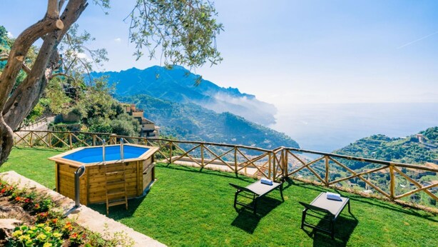 A pool area with a wooden deck, two lounge chairs, and a view of a mountainous coastline. - Casa Meraviglia Ravello (RAVELLO)