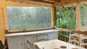 Eine rustikale Küche mit einem großen Fenster, Holzbalken an der Decke, weißen Schränken und einem langen Esstisch mit vier Holz-Untersetzern.