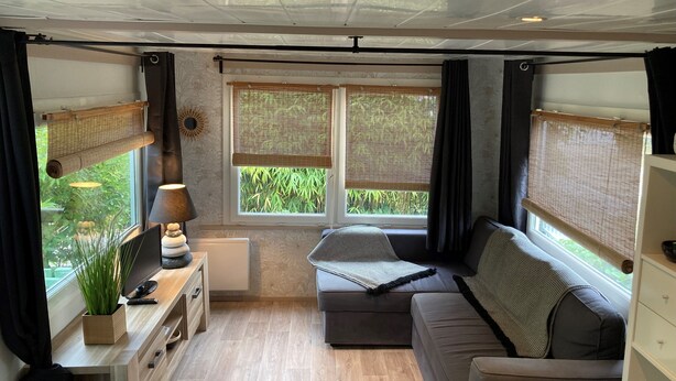 Ein kompakter Wohnraum mit einer Couch, einem kleinen Tisch mit Lampe und einem Fernseher. Zwei Fenster mit Jalousien und Blick ins Grüne.