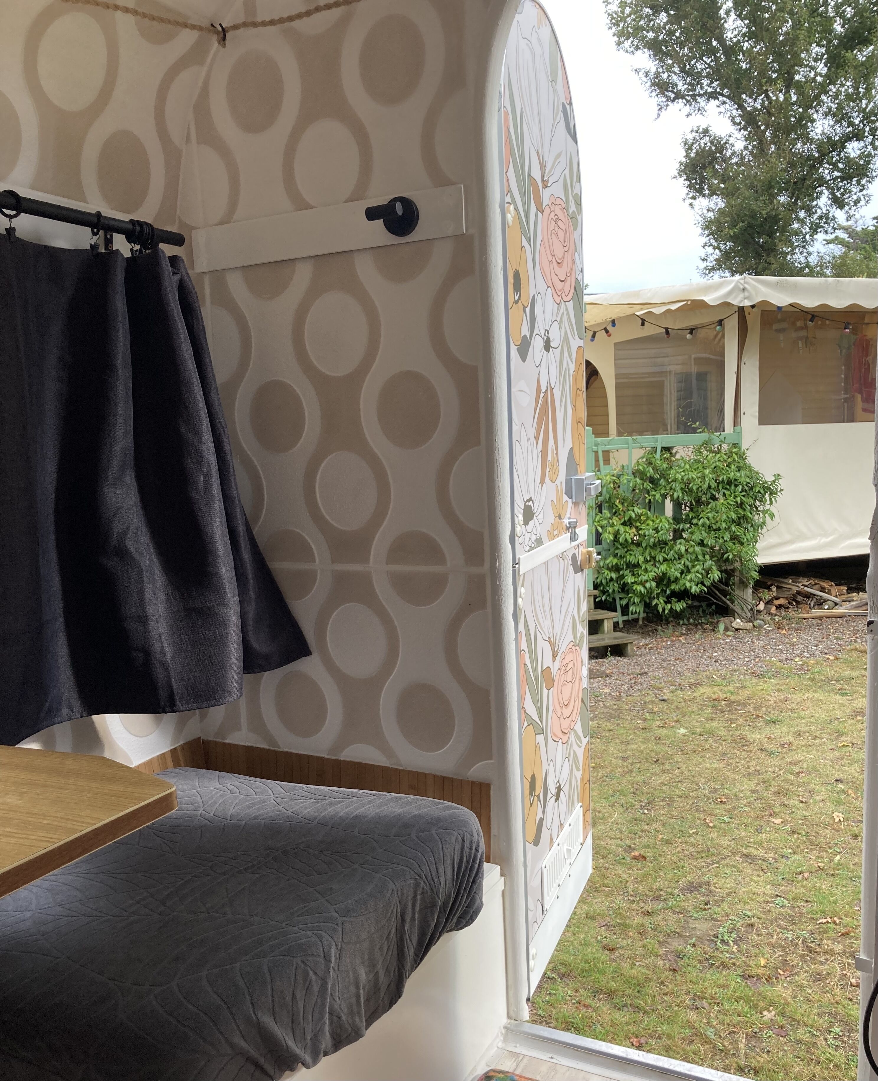 A tent with a bed, a hanging curtain, and a view of a garden with a tree and a building in the background.