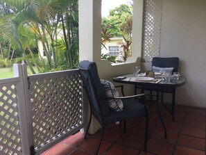 Une terrasse avec une table dressée pour deux, entourée d’une clôture en treillis blanc, donnant sur un jardin luxuriant et un bâtiment.