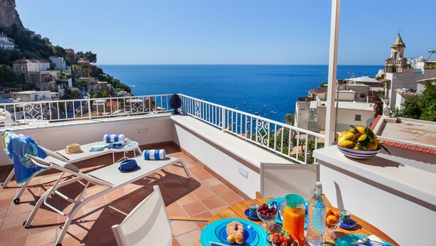 Une terrasse avec une table dressée pour le petit-déjeuner, offrant une vue sur une ville côtière et la mer.