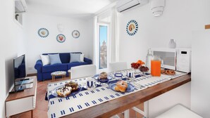 Un salon moderne avec un canapé bleu, une table à manger dressée pour le petit-déjeuner et une vue sur un immeuble par la fenêtre.