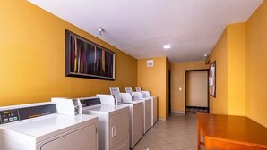 A laundry room with multiple washing machines and dryers, a wooden table, and a large abstract painting on the wall. - Sun Valley Suites (Bullhead City)