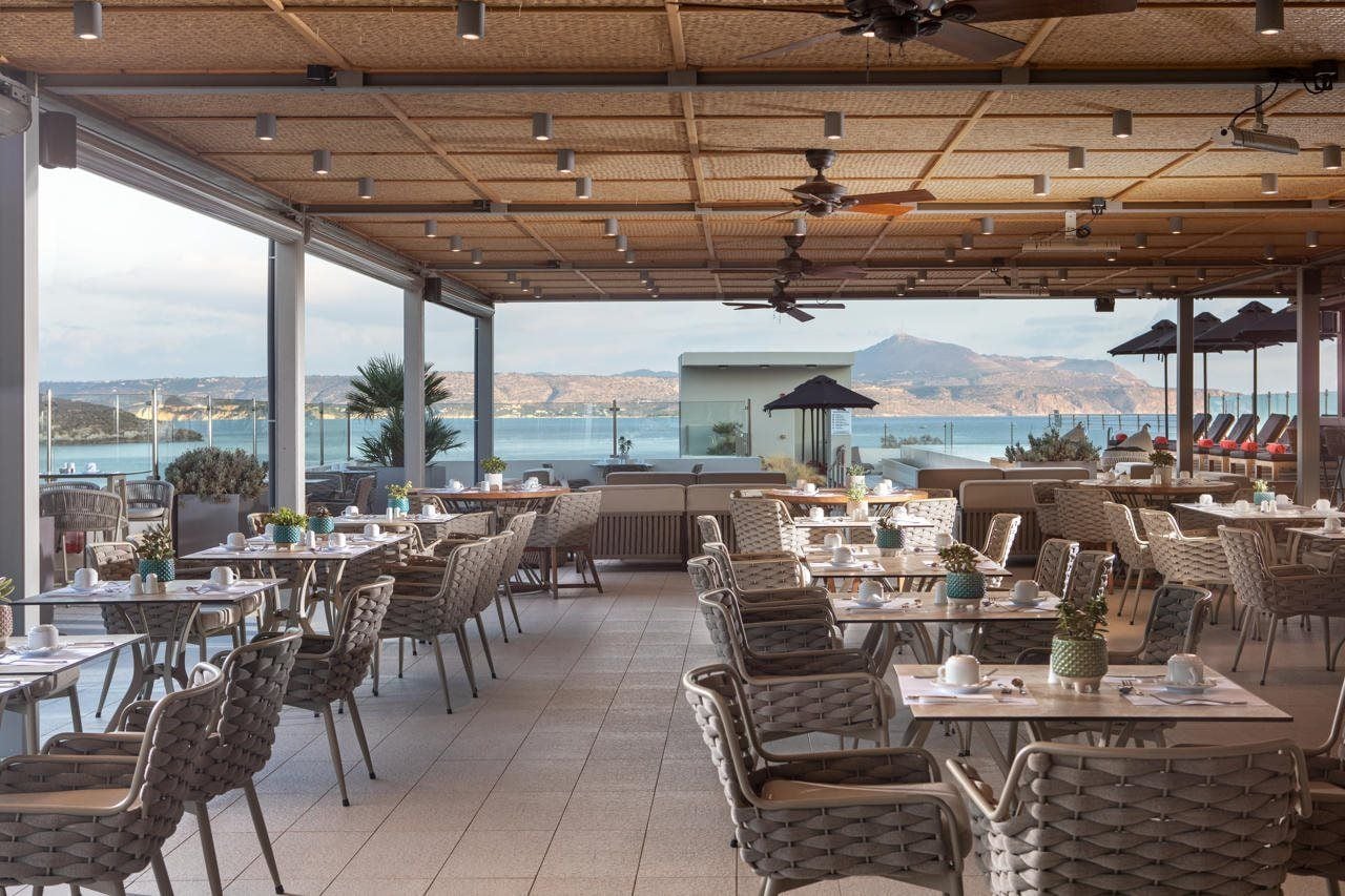 Un espace de restauration en plein air, avec des chaises et des tables en osier, offrant une vue sur un plan d’eau et des collines.