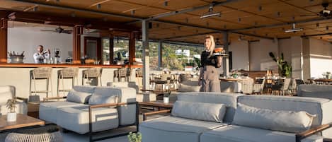 Een terras met zitgedeelte, inclusief banken, stoelen en tafels. Er zijn plafondventilatoren en een bar op de achtergrond.
