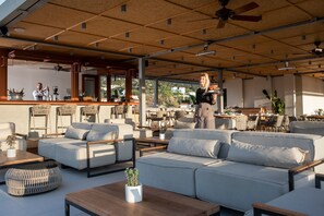 Un espace extérieur aménagé avec des canapés, des fauteuils et des tables. On peut voir des ventilateurs de plafond et un bar au fond.