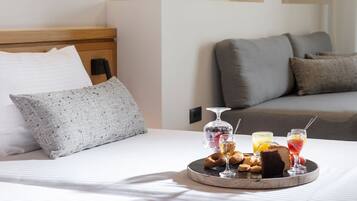 A neatly made bed with a tray of breakfast items including pastries, a glass of juice, and a glass of wine, in a modern hotel room with a grey sofa and a patterned pillow.