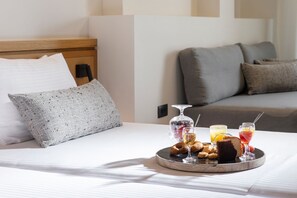 Ein ordentlich bezogenes Bett mit einem Frühstücksbrett, das Gebäck, ein Glas Saft und ein Glas Wein zeigt, in einem modernen Hotelzimmer mit grauem Sofa und einem gemusterten Kissen.