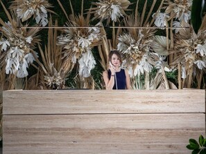 Iemand staat voor een houten bank, met een achtergrond van gedroogde planten en begroeiing.