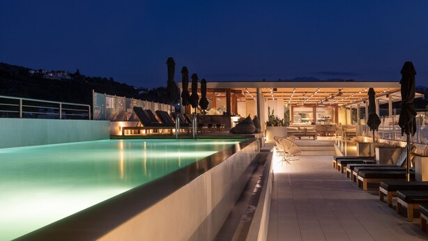 Un espace aménagé au bord de la piscine, avec des chaises longues et des parasols, une piscine, et un bâtiment doté d’une façade en verre.