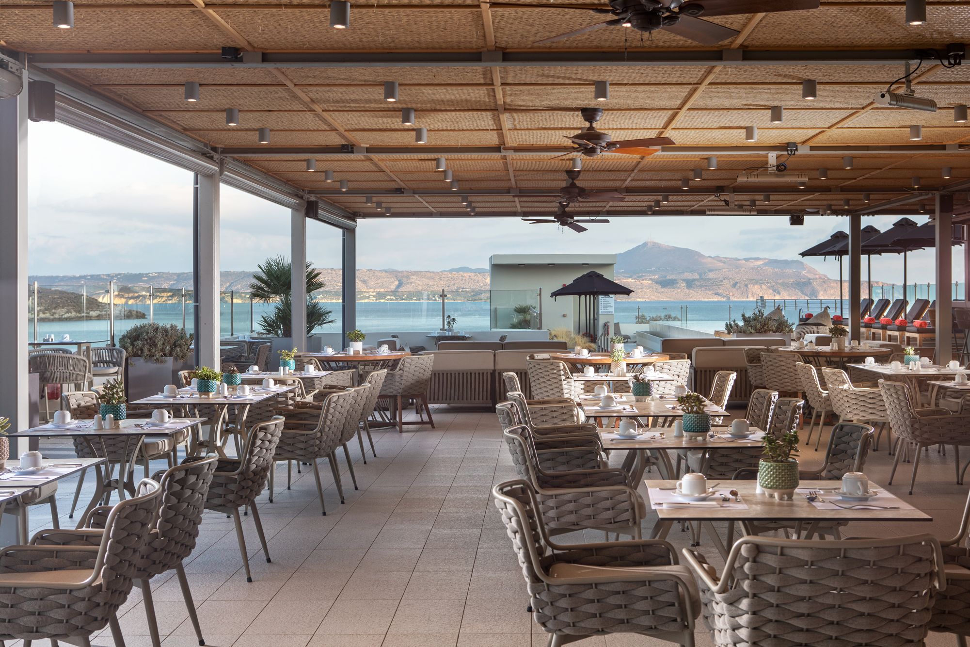 Un área de comedor al aire libre con sillas y mesas de mimbre, ventanas amplias con vistas al mar y las colinas, y ventiladores de techo.