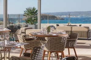Un restaurant en bord de mer, avec des tables rondes et des chaises en osier, offrant une vue sur l’océan.