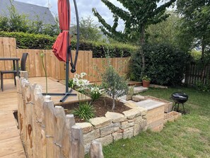 Une terrasse en bois avec une balançoire, un barbecue et un petit jardin agrémenté de plantes et d’un arbre.