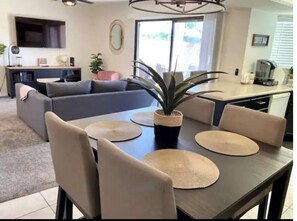 A modern living room with a sofa, dining table with chairs, and a kitchen area in the background. - Casa Del Rio - Across from Rotary Park, River, Boat launch, swimming & fishing (Bullhead City)