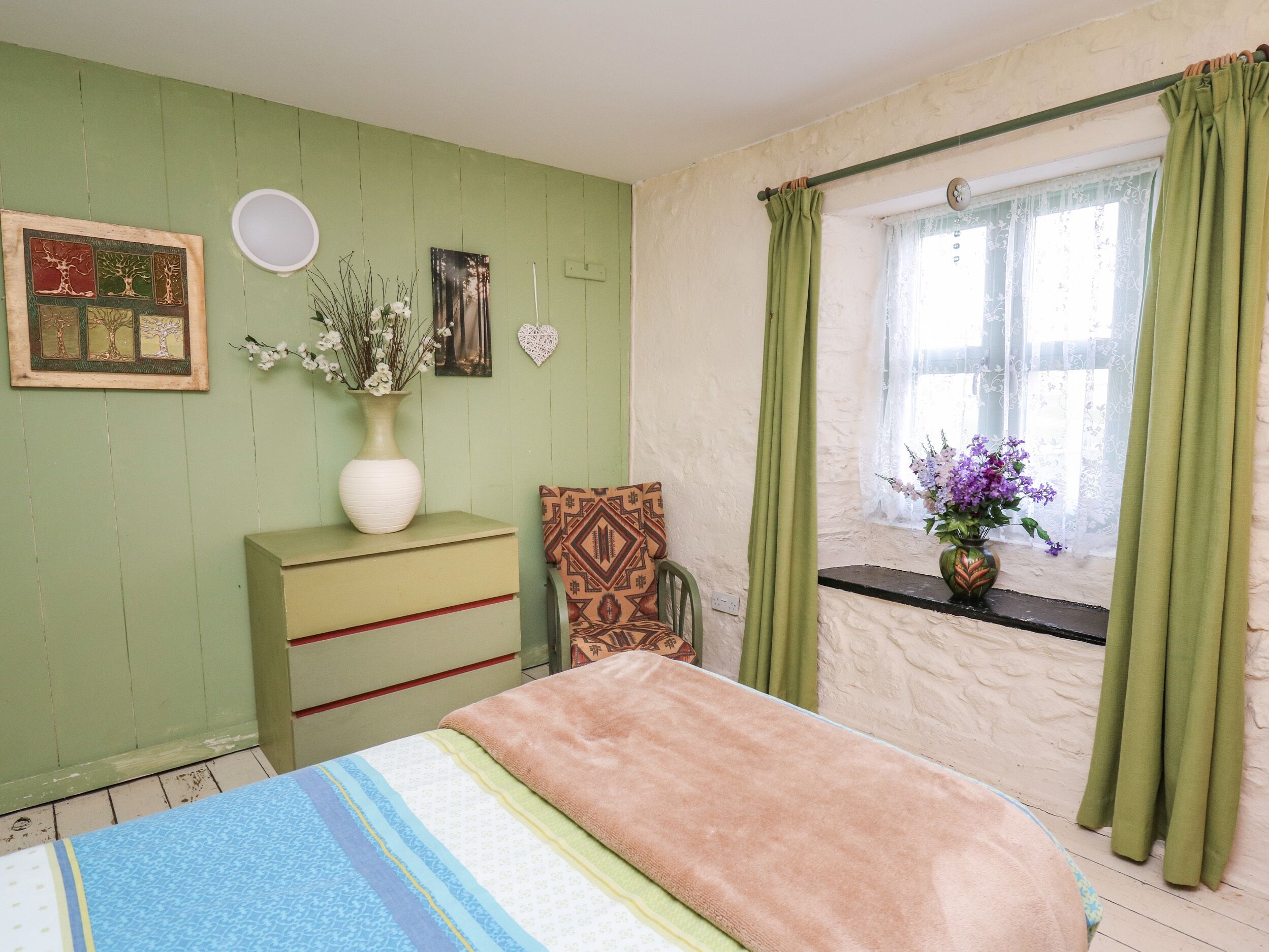 A bedroom with a bed, a dresser, a chair, a window with curtains, and wall decorations including a vase with flowers and framed pictures.