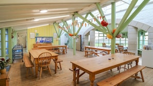 Une pièce spacieuse avec des tables et des bancs en bois, des poutres métalliques vertes et de grandes fenêtres qui laissent entrer la lumière naturelle.