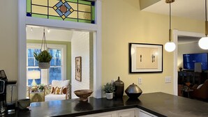 A kitchen with a black countertop, white cabinets, a hanging plant, a framed picture, and a television in the background.