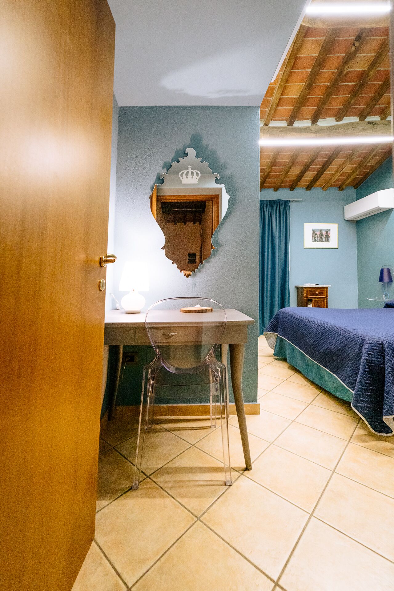 A room with a wooden door, a desk with a chair, a bed with a blue bedspread, a wall-mounted mirror, and a framed picture.