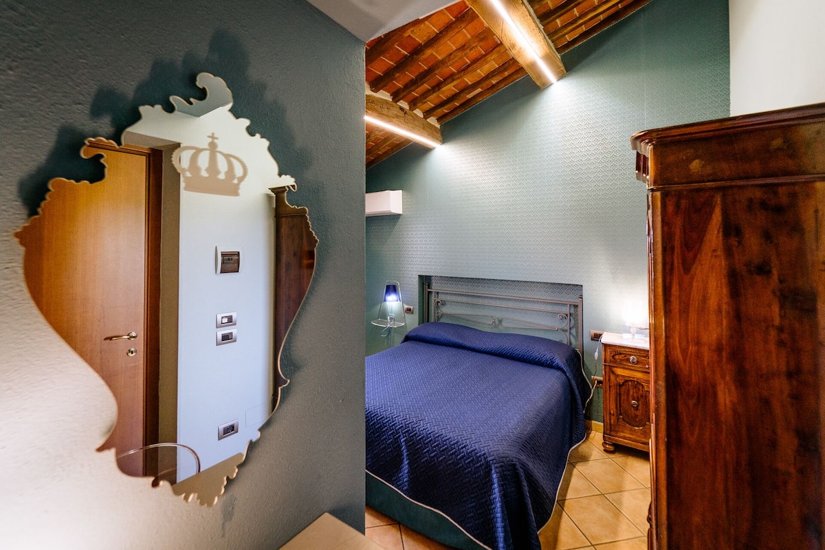 A bedroom with a wooden wardrobe, a bed with a blue blanket, a nightstand, and a wall-mounted lamp.