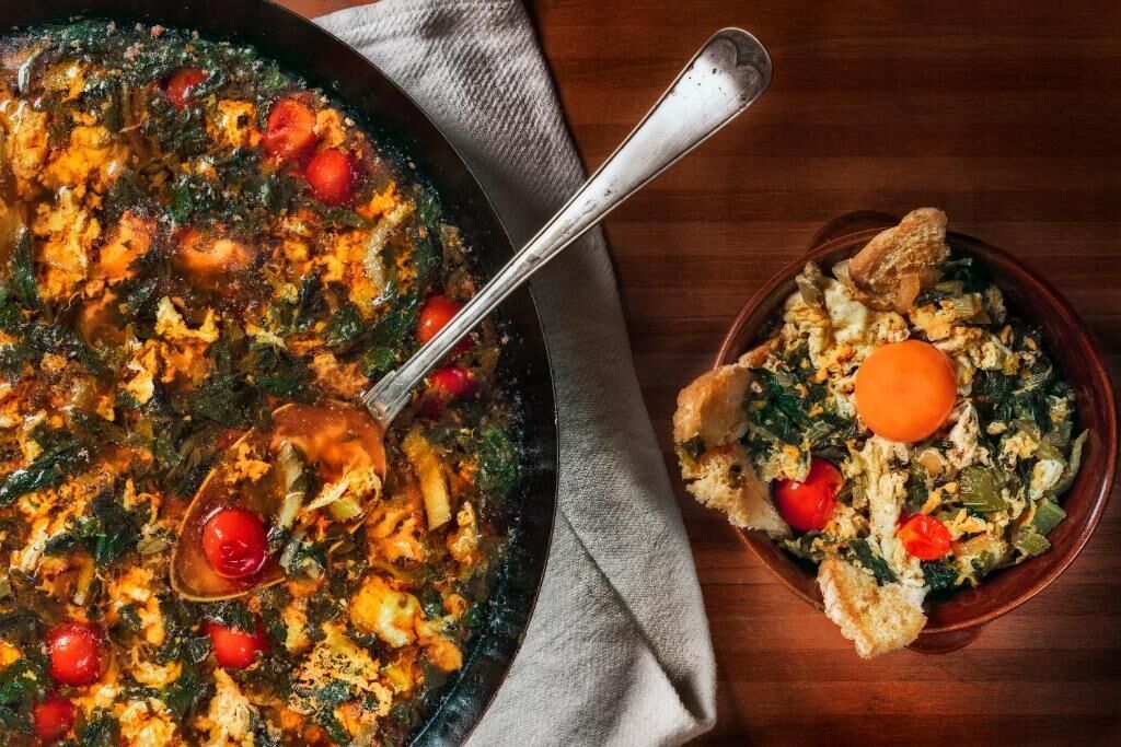 A skillet with a stew containing tomatoes and leafy greens, accompanied by a bowl of scrambled eggs and bread.