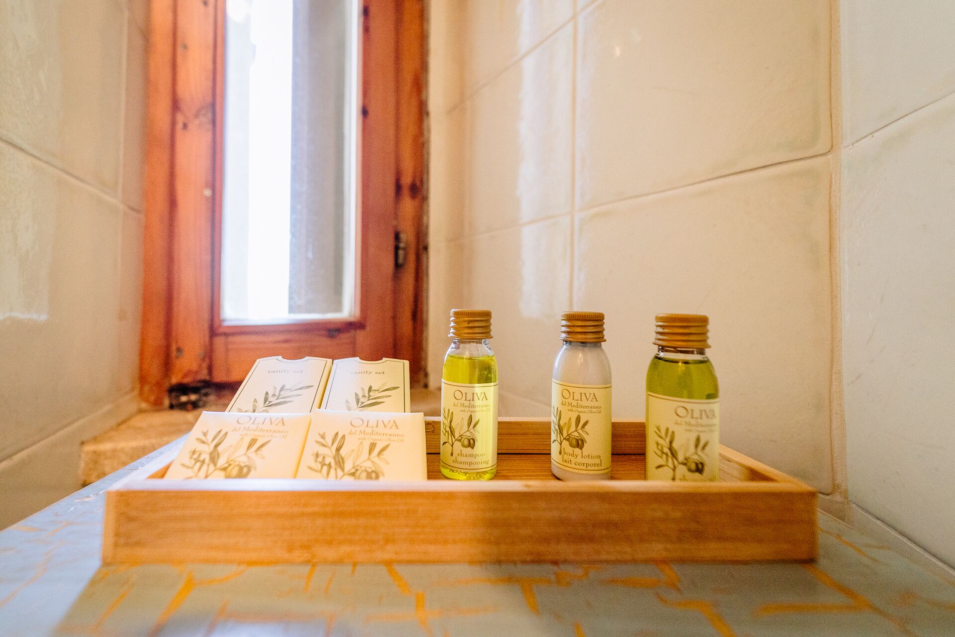 A wooden tray with three bottles of olive oil and a booklet on a tiled floor near a window.