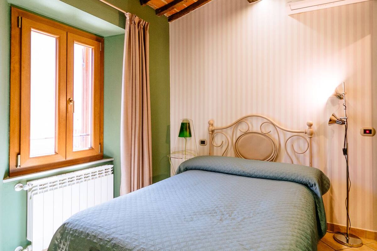 A bedroom with a bed, a wooden ladder, a radiator, a window with curtains, and a wall-mounted lamp.