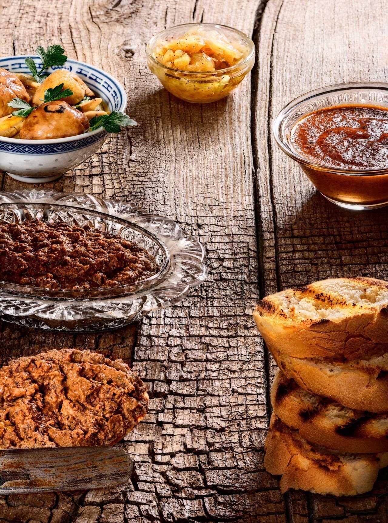 A rustic wooden table with various food items including a bowl of soup, a bowl of fruit, a bowl of sauce, and a stack of bread.