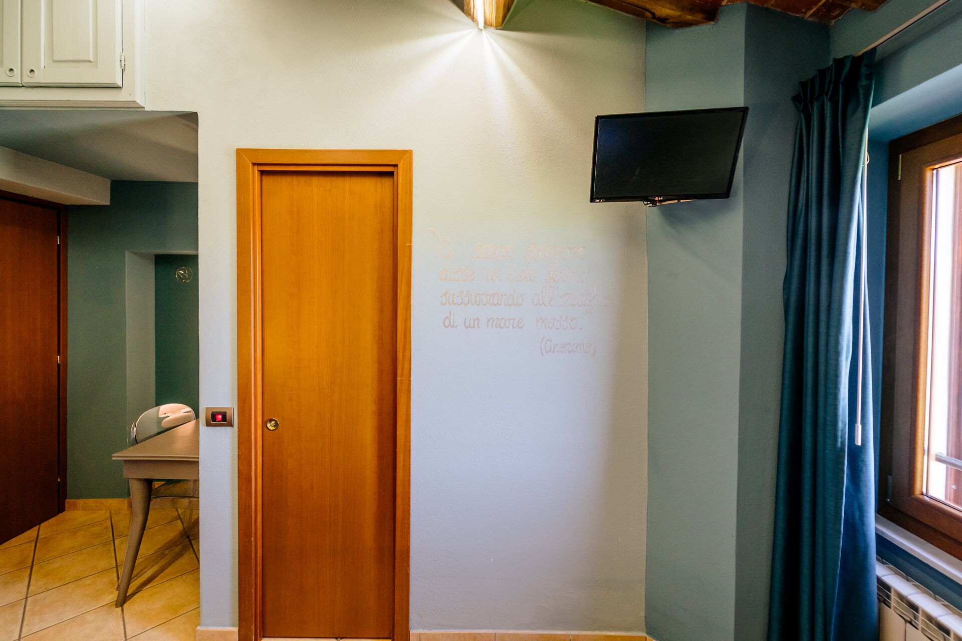 A room with a wooden door, a white wall with a quote, a flat-screen TV, and a window with curtains.