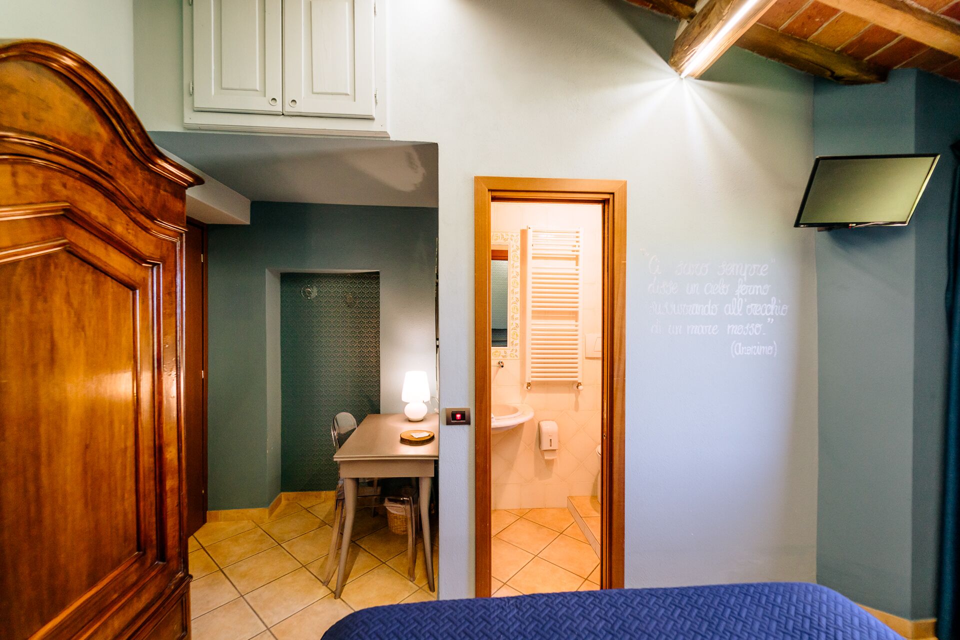 Een slaapkamer met een bed, een bureau met stoel, een houten kledingkast, een badkamer met douche en een tv aan de muur.