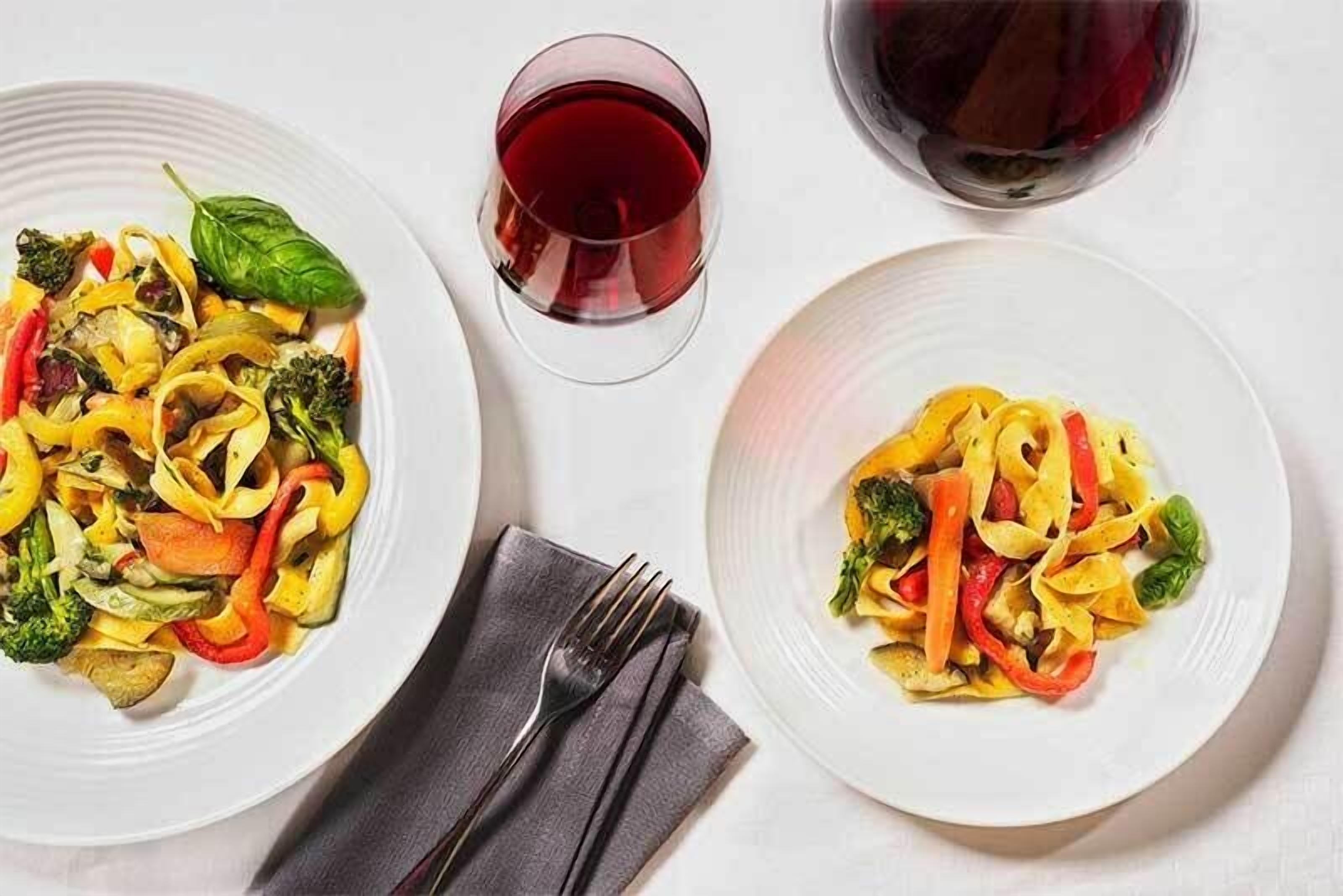 Due piatti di pasta con verdure e un bicchiere di vino rosso su un tavolo bianco.
