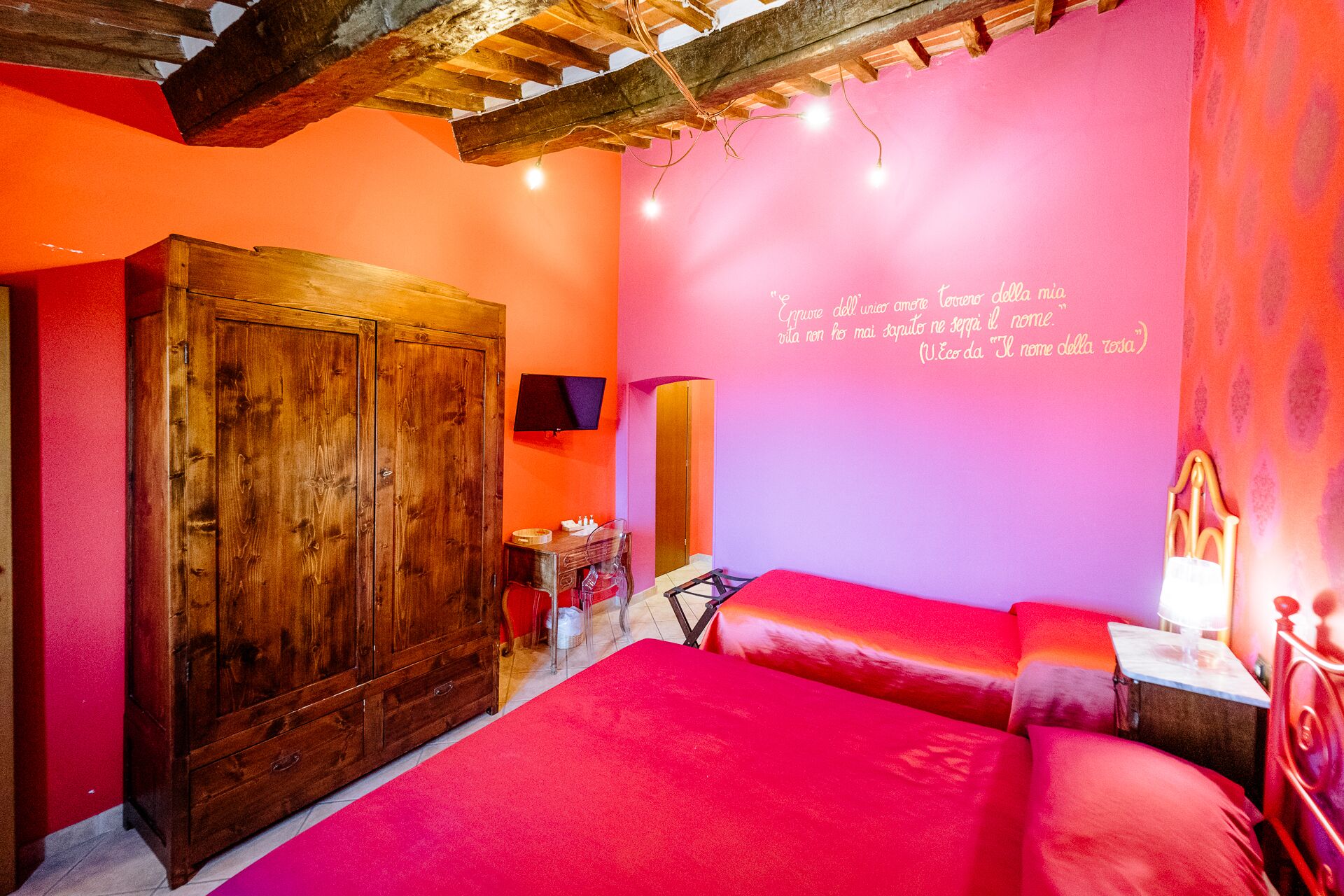 A room with a wooden wardrobe, a bed with red bedding, a wall-mounted TV, and a quote in Italian.