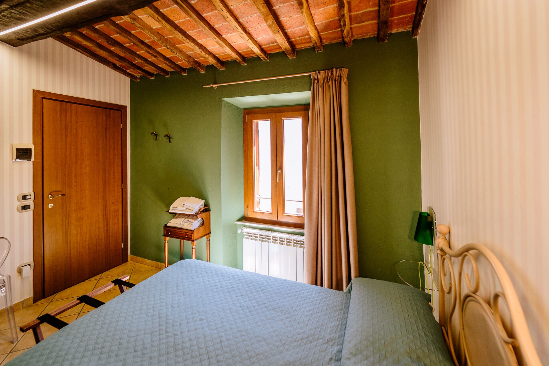 A bedroom with a bed, a wooden door, a window with curtains, and a small table with a lamp.