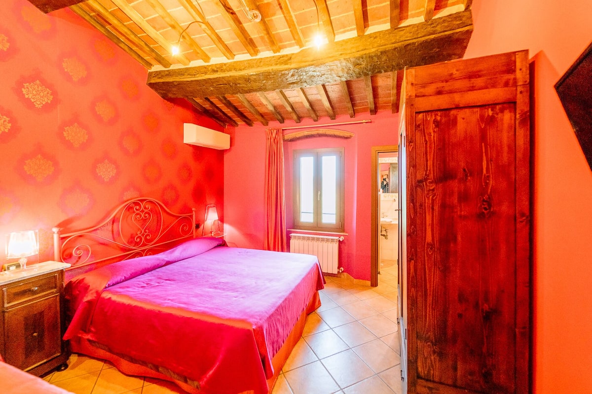 A bedroom with a wooden ceiling, a bed with a pink bedspread, a nightstand, a window, and a door.