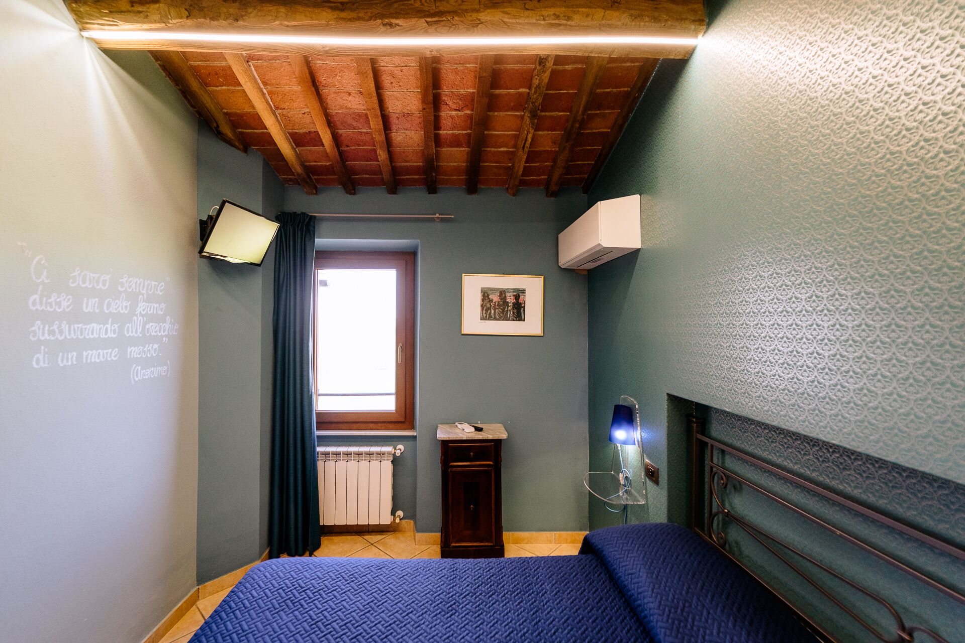 Une chambre avec un plafond en bois, un lit recouvert d’une couverture bleue, un radiateur, une petite armoire, un tableau encadré et un climatiseur fixé au mur.