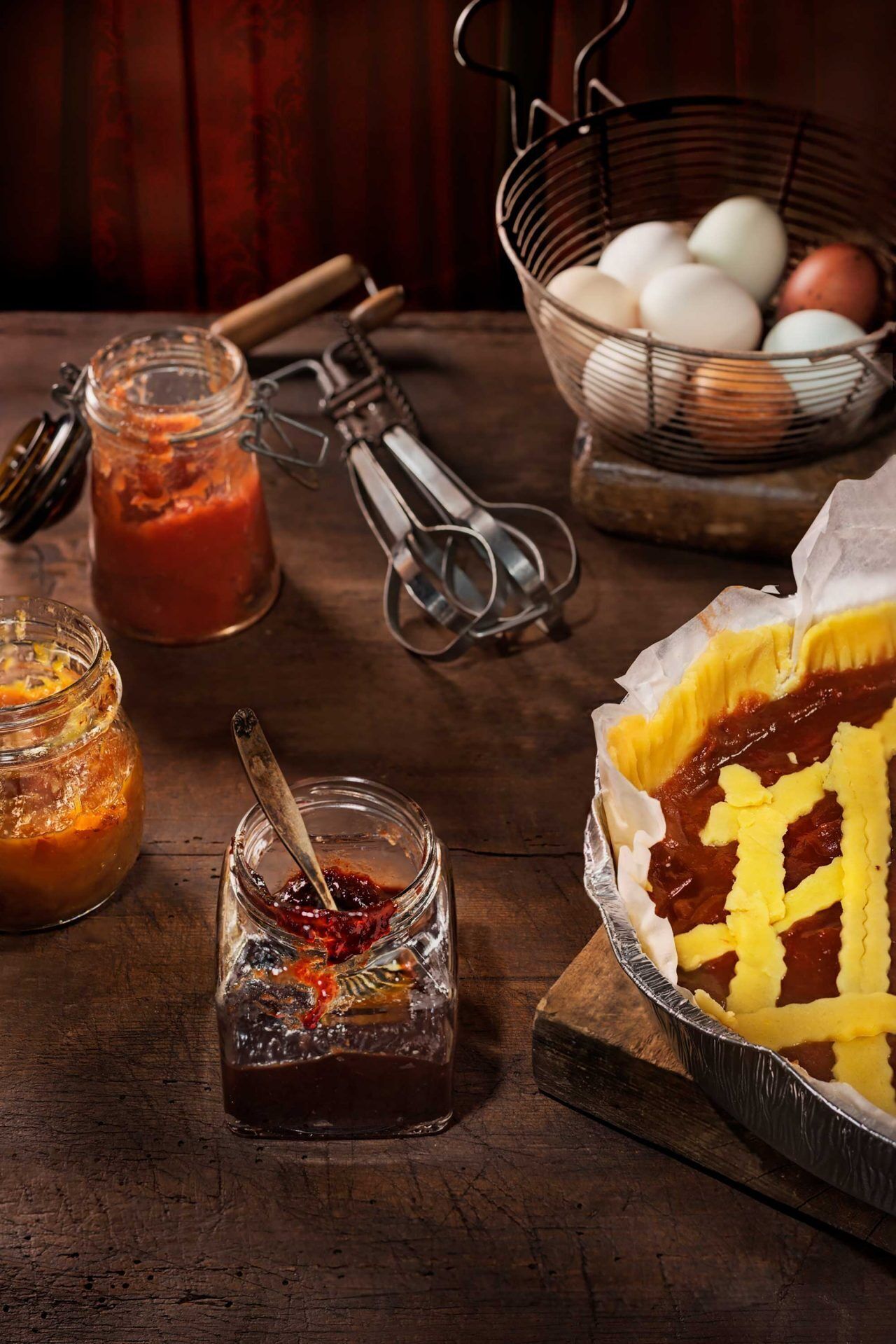 Une cuisine rustique avec une tarte sur un plat recouvert de papier d’aluminium, un pot de confiture et un panier d’œufs.