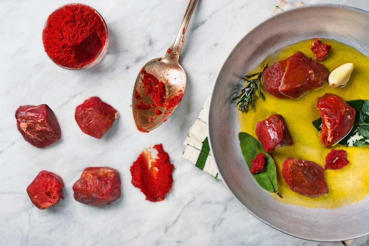 Un bol contenant de la nourriture avec de la viande et des herbes, une cuillère avec une poudre rouge, et des morceaux de viande éparpillés sur une surface en marbre.