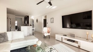 A modern living room with a white sofa, a glass coffee table, a large flat-screen TV, a ceiling fan, and a kitchen in the background. - Modern 4 Bdr Home near Tradition Town Center (Port St. Lucie)