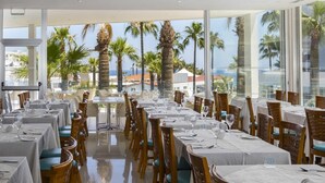 A dining area with tables set for service, large windows with a view of palm trees and buildings, and a reflective floor. - Tsokkos Beach Hotel (Paralimni)