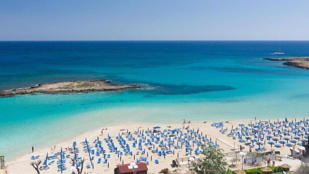 A beach with rows of sun loungers and umbrellas, a clear blue sea, and rocky outcrops. - Tsokkos Beach Hotel (Paralimni)