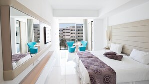 A modern hotel room with a large bed, a sitting area with two chairs, a large mirror, and a view of the cityscape. - Tsokkos Beach Hotel (Paralimni)