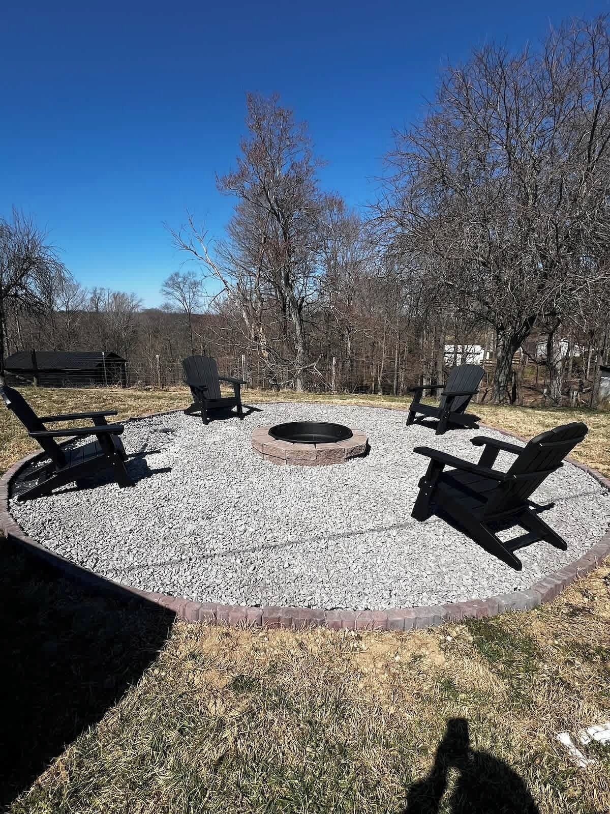 A circular seating area with four Adirondack chairs around a fire pit, surrounded by gravel and trees.