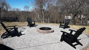 A circular seating area with four Adirondack chairs around a fire pit, surrounded by gravel and trees.