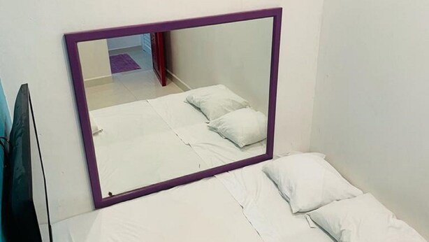 Una cama pequeña y bien hecha con sábanas y almohadas blancas, un espejo con marco morado encima y una pared azul a la izquierda.