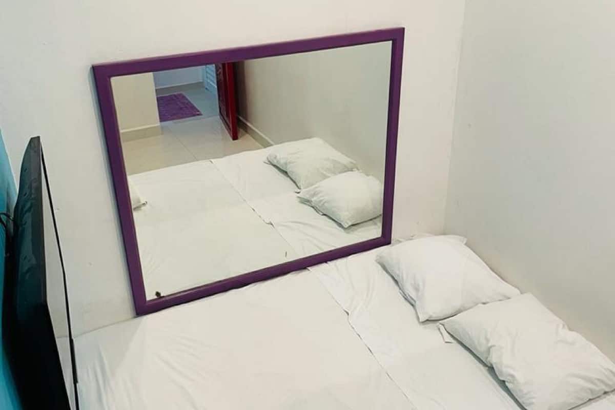 Una cama pequeña y bien hecha con sábanas y almohadas blancas, un espejo con marco morado encima y una pared azul a la izquierda.