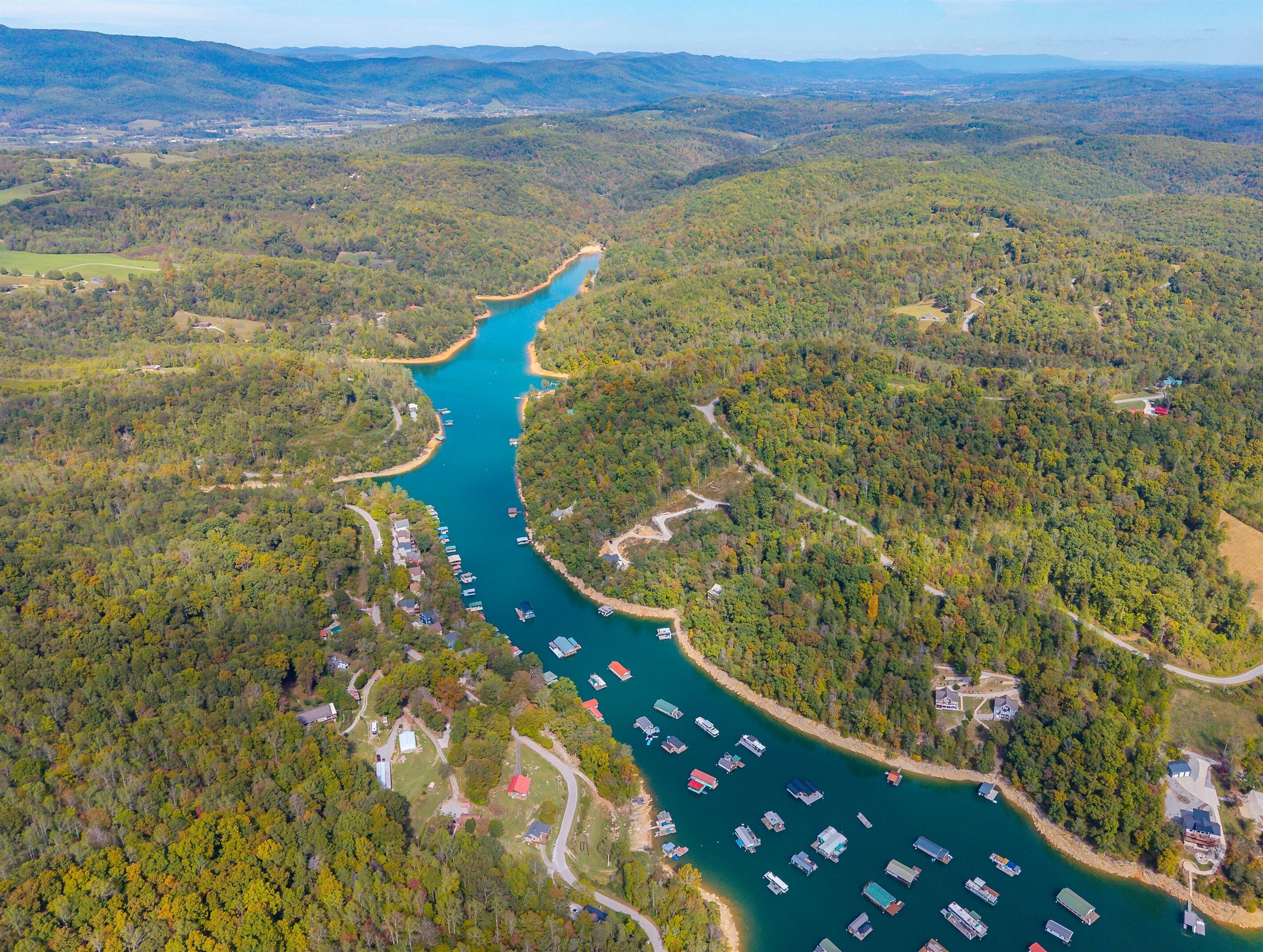 Une vue aérienne d’une rivière où des bateaux sont amarrés le long de ses rives, le tout étant entouré d’une forêt dense et de zones résidentielles.