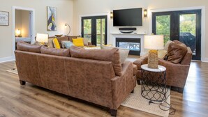 A spacious living room with a large brown leather sofa, a smaller brown armchair, a fireplace, a flat-screen TV, and a large window with a view of trees. - 25% late summer discount! Lakefront | 2 slip covered dock | Hot tub | (Speedwell)