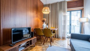 A modern hotel room with a wooden wall, a flat-screen TV, a dining table with chairs, a sofa, and a large window with curtains. - Firmeza388 2E- Sober Apartment by LovelyStay (Porto)