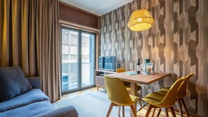 Ein modernes Hotelzimmer mit einer Couch, einem Esstisch mit Stühlen, einem Fernseher und einem großen Fenster mit Vorhängen.