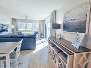 A modern living room with a sofa, dining table, and chairs, a large window with a view, and a ceiling fan.